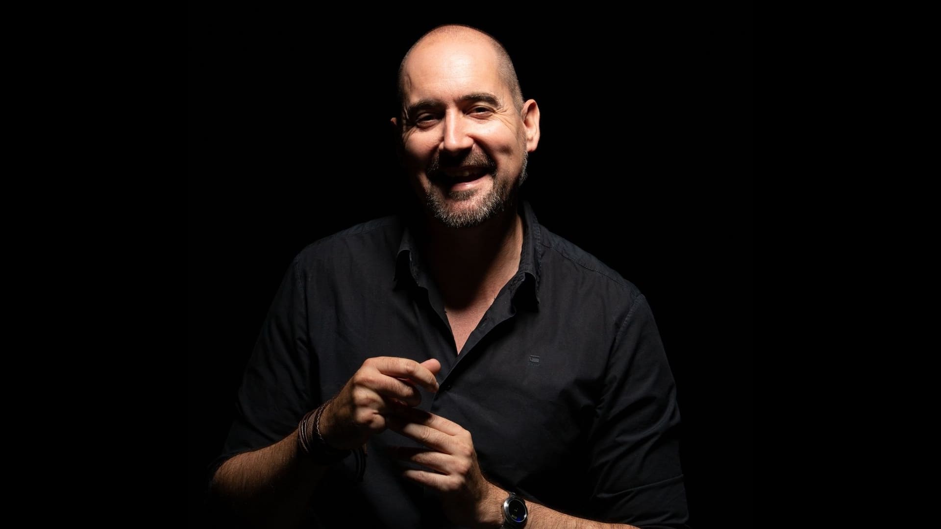 Huw Davies laughing, hands raised mid-gesture, lit against a black background.